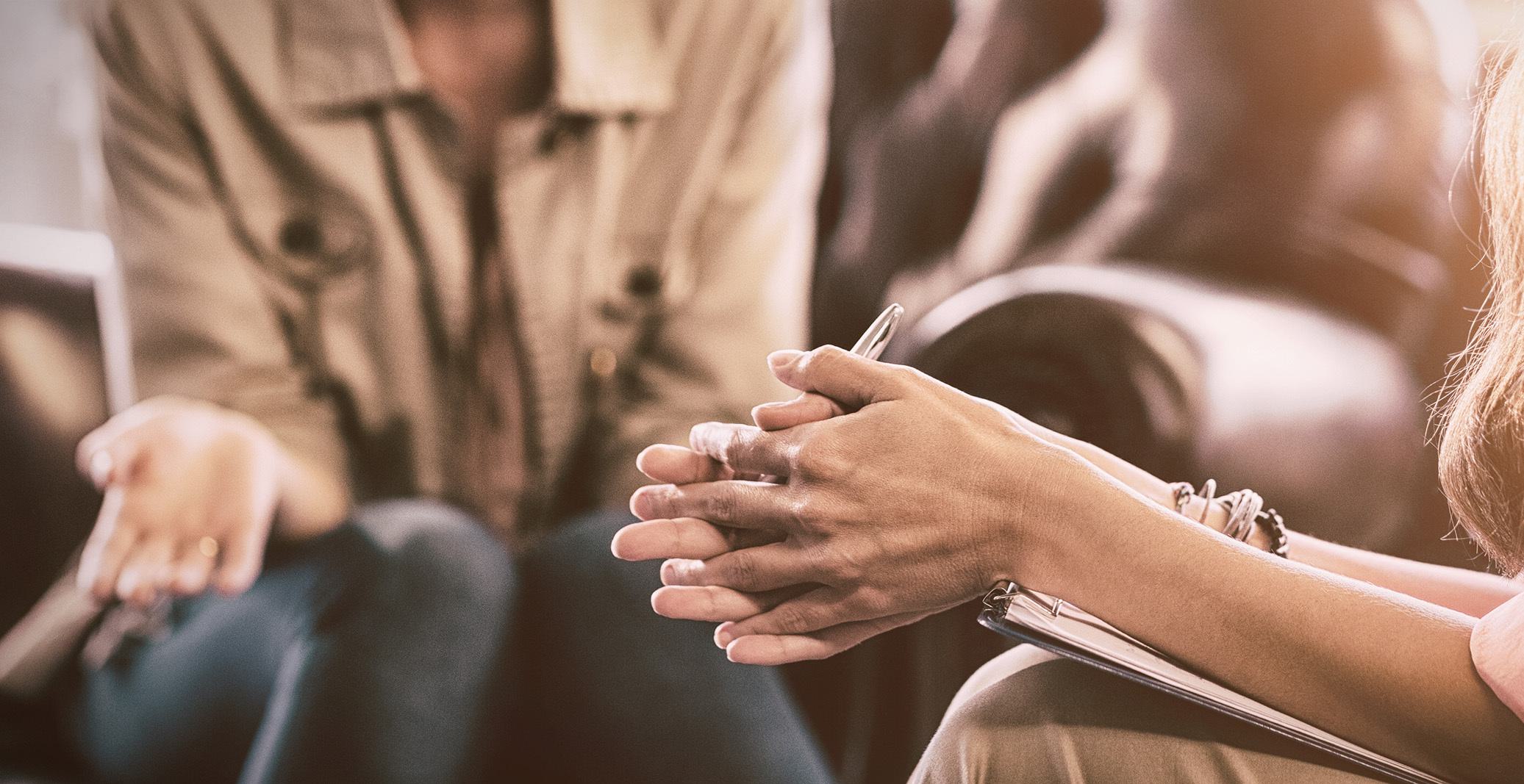 a seated female psychotherapist with clasped hands talking to another person