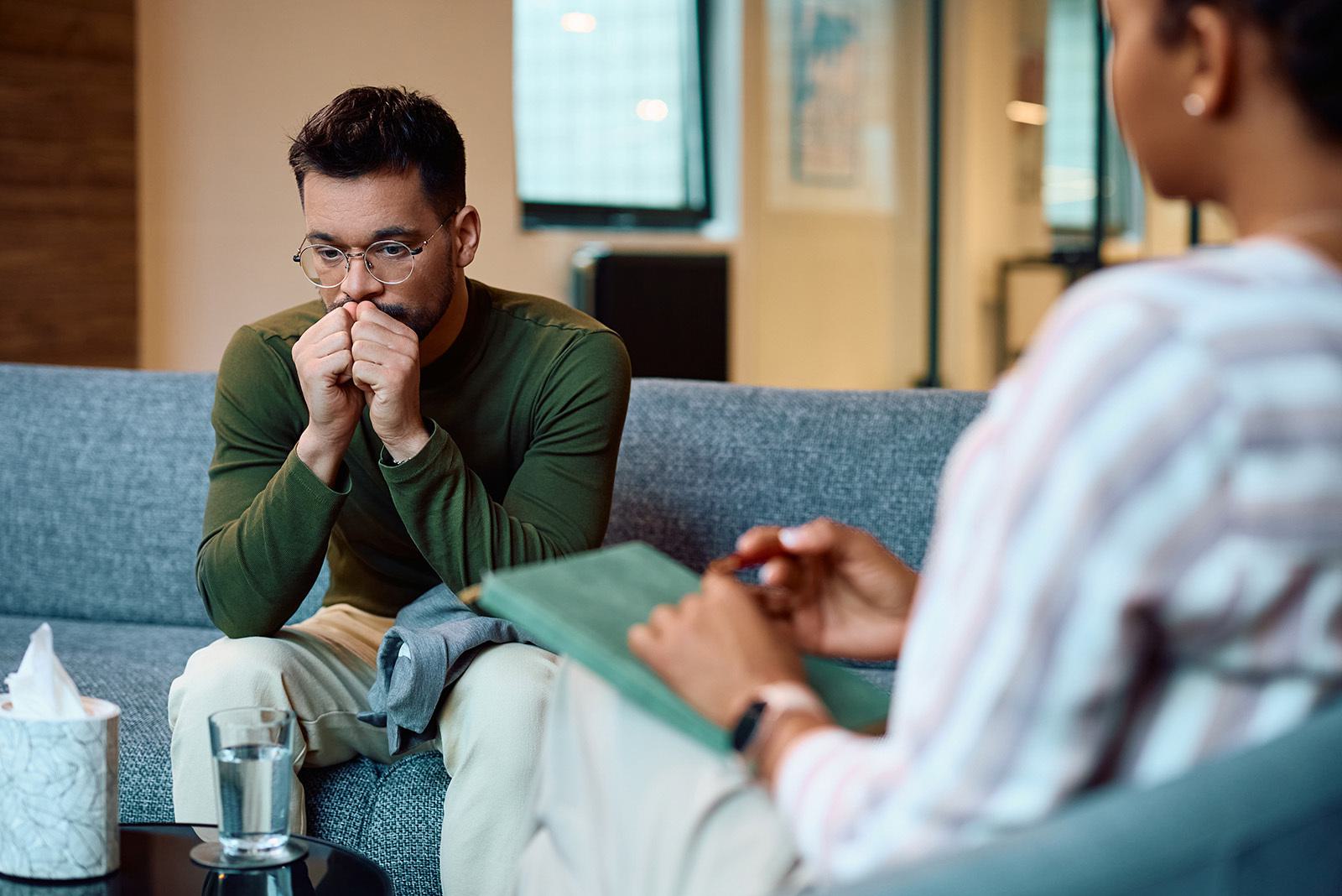 a young man looking thoughtful sitting on a sofa talking to a pychotherapist