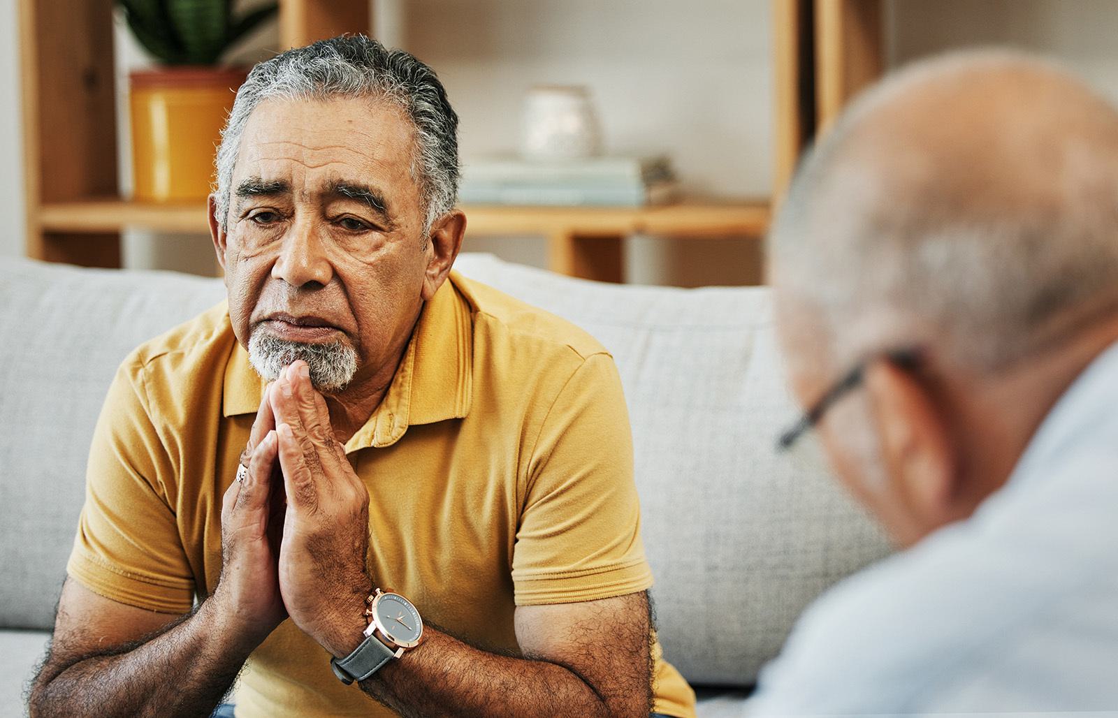 a male patient in a psychotherapy session sitting on a sofa with his hands clasped to his chest whilst looking concerned