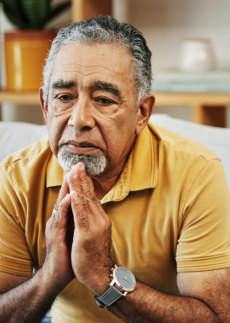 a male patient in a psychotherapy session sitting on a sofa with his hands clasped to his chest whilst looking concerned